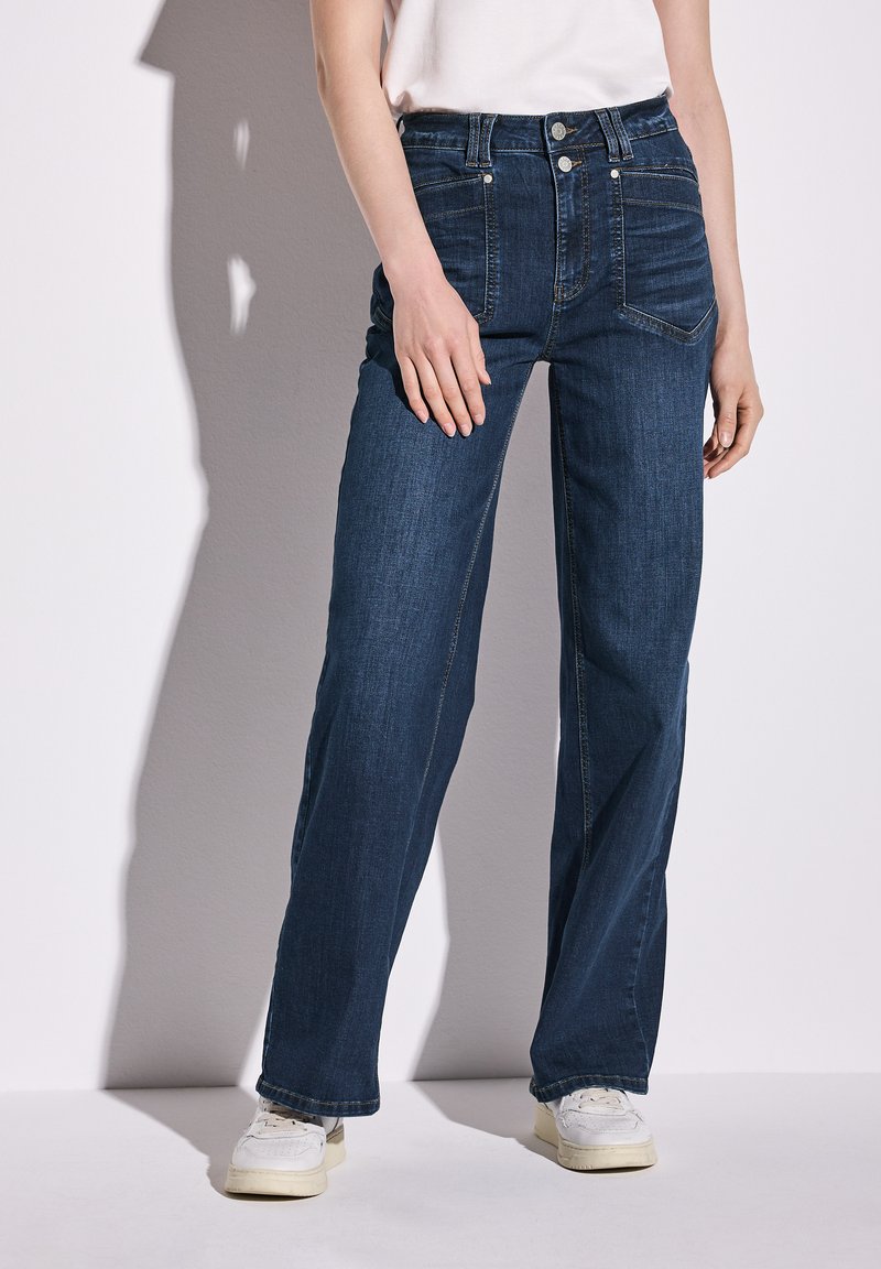 Jeans de tiro alto en azul oscuro con un diseño de pierna ancha, que cuentan con bolsillos frontales y un cierre de botones, combinados con zapatillas blancas.