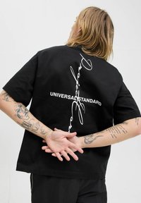 Sort t-shirt med korte ærmer lavet af bomuld, med hvid tekst "UNIVERSAL STANDARD" og et kuperet design på ryggen. Casual pasform.