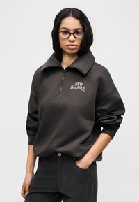 Σκούρο γκρι φερμουάρ φούτερ με ψηλό γιακά. Λογότυπο "NEW BALANCE" σε λευκό, ριμπ μανσέτες και άνετη εφαρμογή. Φοριέται πάνω από σκούρο παντελόνι.