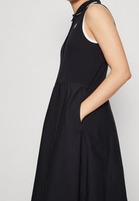 Czarna sukienka bez rękawów z kołnierzykiem i bocznymi kieszeniami, noszona przez osobę, która trzyma jedną rękę w kieszeni na tle jednolitym.