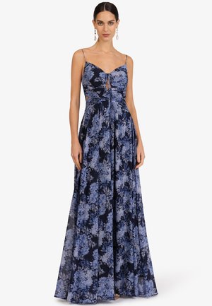 Frau trägt ein ärmelloses, marineblaues Maxikleid mit hellblauem Blumenmuster und dünnen Trägern, steht vor einem weißen Hintergrund.