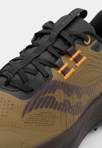 Chaussure de course présentant un dessus noir et marron, des matériaux texturés, des accents orange et violet, ainsi qu'un système de laçage solide.