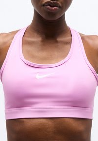 Κοντινό πλάνο ενός ατόμου που φοράει αθλητικό σουτιέν Nike σε ανοιχτό ροζ χρώμα, που δείχνει τον λαιμό, τους ώμους και το πάνω μέρος του στήθους με καθαρό λευκό φόντο.