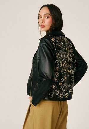 Mujer con chaqueta de cuero negra con tachuelas doradas y plateadas que forman patrones florales en la espalda, combinada con pantalones beige.