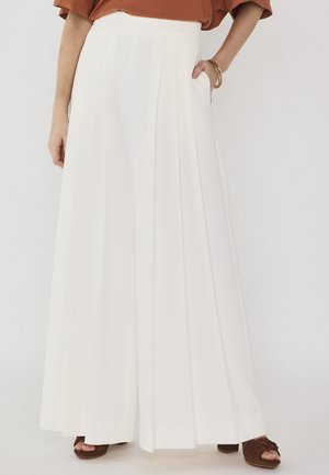 Donna con gonna lunga plissettata bianca, top marrone, braccialetti dorati e sandali platform marroni con punta aperta, in piedi con una mano in tasca.