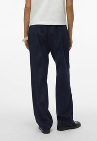 Person som bär marinblå randiga vida byxor, vit topp, svarta loafers, sedd bakifrån mot en helt vit bakgrund.