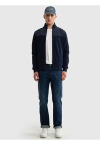 Man met een blauwe baseballpet, een marineblauwe jas met rits, een wit T-shirt, opgerolde spijkerbroek en witte sneakers, staand tegen een effen achtergrond.