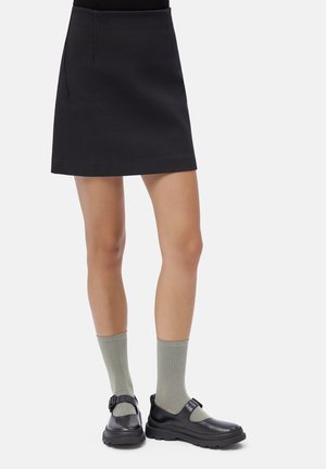 Donna con una mini gonna nera, calze a coste grigio chiaro e scarpe in pelle nera con fascetta contro uno sfondo bianco.