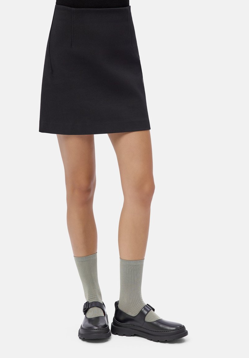 Vrouw in een zwarte mini-rok, lichtgrijze geribbelde sokken en zwarte leren schoenen met gespsluiting tegen een witte achtergrond.