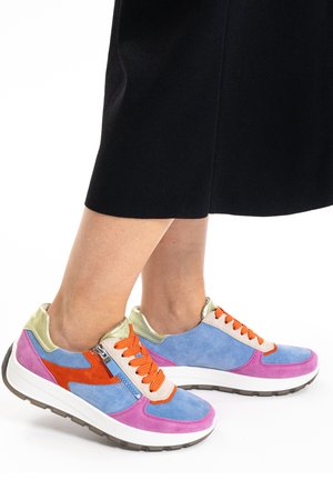 Piernas con zapatillas multicolores con detalles en azul, rosa, naranja, beige y dorado, combinadas con pantalones negros recortados.