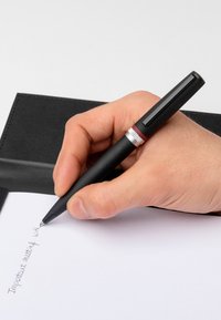 Schwarzer Stift mit silberner und roter Akzent, der über weißem Papier gehalten wird, schreibt "Wichtige Besprechung mit" neben einem schwarzen Notizbuch.