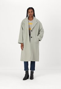 Hellgrauer übergroßer Mantel mit Reverskragen, Knöpfen vorne und seitlichen Taschen, kombiniert mit einem gestreiften Pullover und dunklen Jeans.