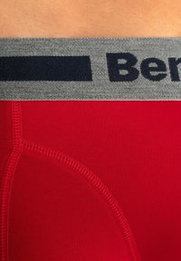 Red boxer briefs with a grey elastic waistband featuring a navy brand logo. Flat seams for comfort and a smooth, stretch fabric texture.
