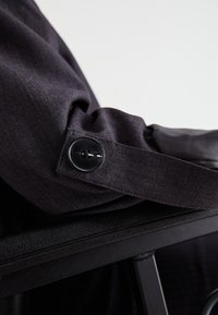 Σκούρος ύφασμα με διακριτή υφή, που διαθέτει κλείσιμο με μαύρο κουμπί στον καρπό. Το κουμπί έχει σχεδίαση με δύο τρυπές, τονισμένο από καθαρές γραμμές.