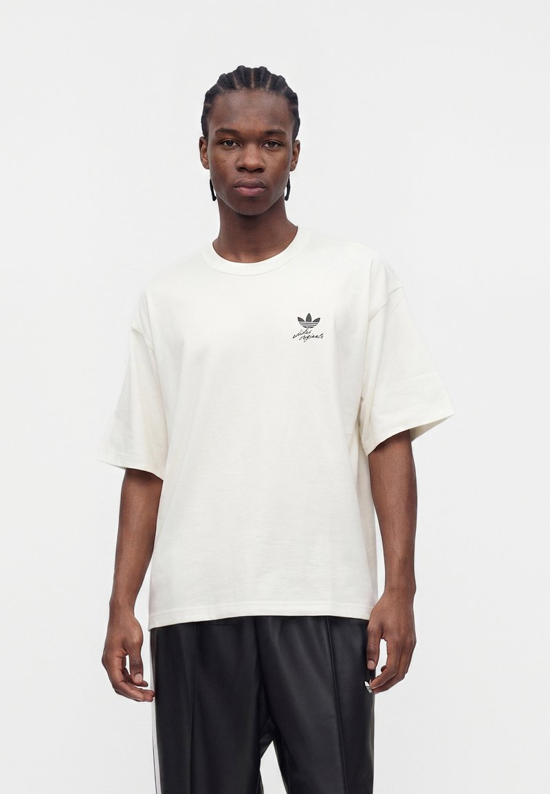 Jeune homme aux cheveux tressés portant un t-shirt blanc ample Adidas Originals et un pantalon de survêtement noir Adidas, debout devant un fond uni.