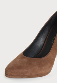Gros plan d'une chaussure à talon haut en suède marron à bout pointu avec une doublure intérieure noire et une semelle plateforme légère.