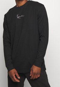 Pitkähihainen musta puuvillainen t-paita, jossa valkoinen allekirjoituslogo rinnassa, pyöreä pääntie ja istuvat hihansuut.