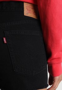 Pantalones cortos de mezclilla negra con un parche de cuero de la marca Levi's, costuras visibles en los bolsillos traseros y un dobladillo deshilachado. Manga de un sudadera roja cercana.
