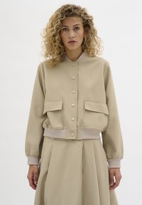 Tânără cu păr ondulat blond îmbrăcând o jachetă bomber bej cu butoni și o fustă plisată asortată, stând în fața unui fundal alb simplu.