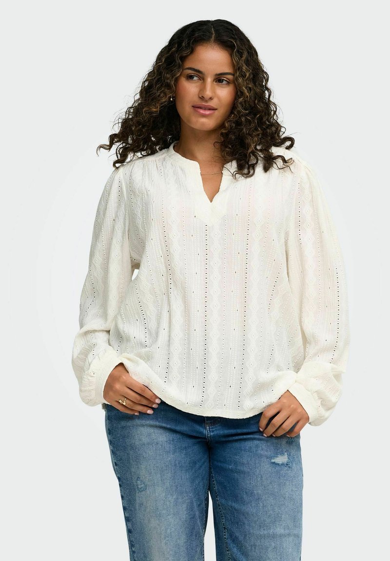 Femme aux cheveux bouclés portant un chemisier blanc texturé à manches longues et un jean bleu, debout avec les mains posées sur les hanches.