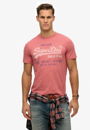 Red heathered short-sleeve t-shirt with white and blue graphic text. Worn with blue jeans and a plaid shirt tied at the waist. Brown watch.