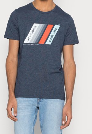 Homme portant un t-shirt bleu marine à manches courtes avec un motif rayé géométrique et les textes "JACK & JONES" et "CORE 1990" sur la poitrine.