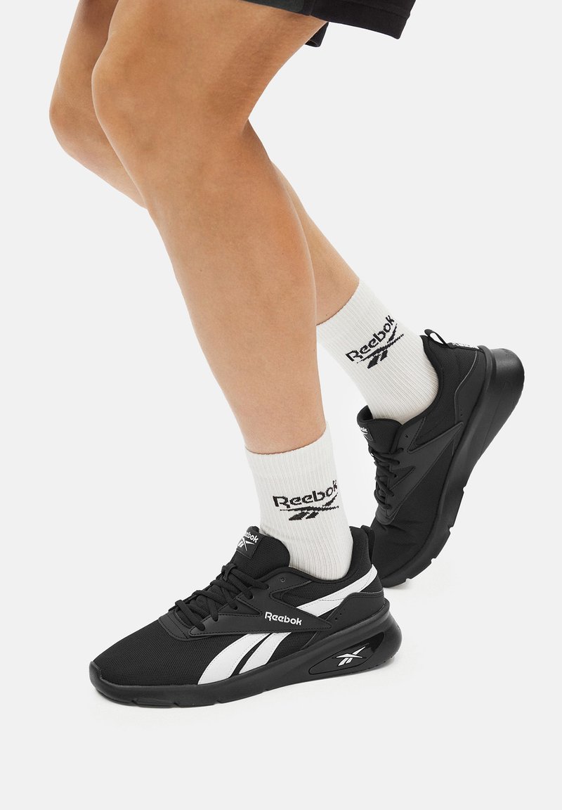 Čierne tenisky Reebok s bielymi detailmi, sieťovinovým zvrškom a plochými šnúrkami, skombinované s bielymi ponožkami s čiernym logom.