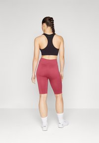 Crni sportski grudnjak s dizajnom racerback, uparen s biciklističkim hlačama boje maroon do koljena. Model nosi bijele čarape do gležnjeva i tenisice.