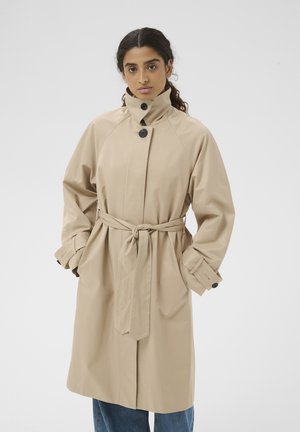 Kvinde iført en beige trenchcoat med sorte knapper og et bundet bælte, stående med hænderne i lommerne mod en ensfarvet baggrund.