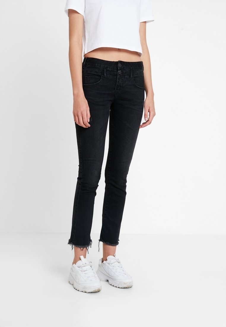 Jeans en denim noir avec une coupe slim, une longueur raccourcie, un ourlet effiloché et un design à cinq poches, associés à des baskets blanches et un top blanc court.