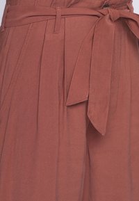 Pantalons couleur rouille avec une ceinture en tissu, dotés de plis et de coutures soignées. Matériau léger avec une texture douce.