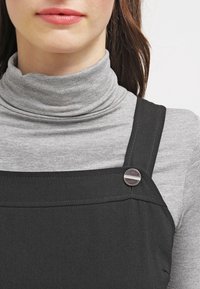 Kvinna som bär en grå polo under en svart ärmlös klänning med en rund metallknapp på axelbandet.