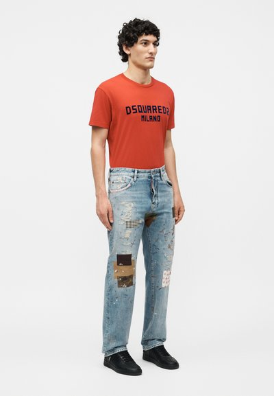 Άνδρας όρθιος φορώντας κόκκινο μπλουζάκι "DSQUARED2 MILANO", ανοιχτό μπλε τζιν παντελόνι με επιδιορθώσεις από ύφασμα και μαύρα αθλητικά παπούτσια.