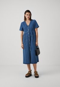 Denim skjortekjole med korte ermer, V-hals, to frontlommer og innsnørt midje, sammen med brune sandal med stropper og en liten svart veske.