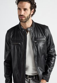 Schwarze Lederjacke mit Stehkragen, zwei Reißverschlusstaschen auf der Brust und glatter Textur. Trägt man über einem weißen Mesh-Oberteil, das ein lässiges Design zur Schau stellt.