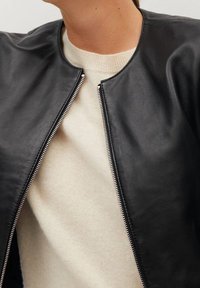Zwarte leren jas met een ronde halslijn, zilveren ritsluiting en zachte interieurstof van een trui zichtbaar eronder.