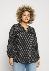 Schwarze Bluse mit weißem geometrischem Muster, Ausschnitt, langen Ärmeln und lockerer Passform, kombiniert mit hellblauen Jeans.