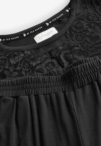 Fekete ruhadarab csipkés virágmintával a nyakkivágás körül és rugalmas derékpánttal, "b by Ted Baker London" felirattal.