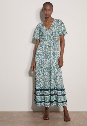 Kobieta w białej i turkusowej sukience maxi w wzór paisley, z krótkimi rękawami, dekoltem w serek i warstwową spódnicą, stojąca na tle jednolitej ściany.