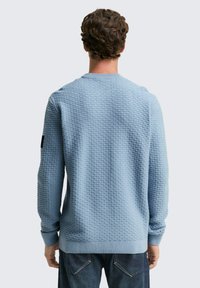 Lichtblauwe gestructureerde trui met een ronde hals, ribgebreide mouwen en zoom. Heeft een subtiel patroon en een zwarte accent op de mouw.