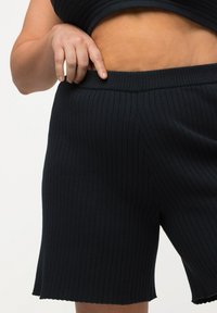 Mustat ribbausneulotut shortsit, joissa on tiukka vyötärönauha, aaltomaisesti viimeistelty helma ja pehmeä pinta, esitettynä lähikuvassa vyötärön alueelta.