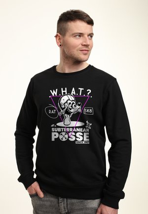 Mężczyzna w czarnej bluzie z karykaturalną szczurzą w hełmie i napisem „W.H.A.T.? Subterranean Posse Since 1967,” patrzący w prawo.