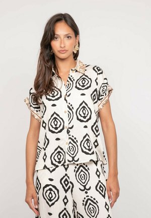 Femme aux longs cheveux bruns portant une chemise blanche à boutons et un pantalon avec des motifs géométriques abstraits noirs, ainsi que des boucles d'oreilles dorées.