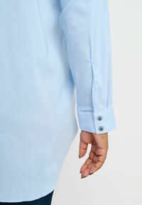 Light blue long-sleeve shirt features a smooth fabric, buttoned cuffs, and a straight hem, with a visible seam along the sleeve.