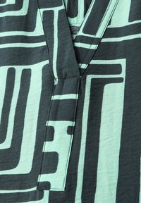 Kurzarmhemd mit V-Ausschnitt. Es hat ein geometrisches Muster in Schwarz und hellem Mint auf einem strukturierten Stoff. Die kräftigen Linien schaffen Kontraste.