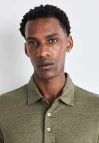 Olivgrön polo skjorta med krage, med en knappslå med tre knappar och strukturerad tyg. Modellen har kort, lockigt hår och en diskret örhänge.