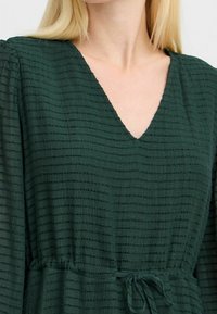 Tumma vihreä mekko, jossa on teksturoitu, ribattu kangas. Siinä on V-pääntie ja sidottu vyötäröyksityiskohta, joka luo istuvan siluetin.