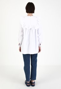 Weißes Blusenshirt mit gerüschtem Rücken, langen Ärmeln mit Schlitzen an den Manschetten, kombiniert mit blauen Jeans und dunklen Sandalen. Einfaches und minimalistisches Design.