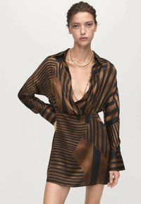 Vrouw in een bruin en zwart gestreepte lange jurk met diepe V-hals en gouden kettingen, poseert met één hand op de heup.