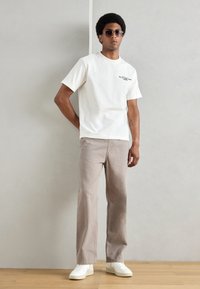Witte katoenen t-shirt met zwarte tekst, gecombineerd met beige katoenen broek. Model draagt donkere zonnebrillen en witte sneakers. Eenvoudig, ontspannen ontwerp.